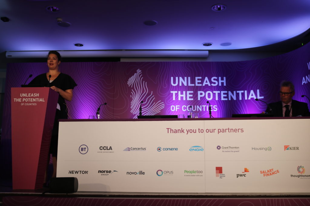 Woman speaking at podium on stage with "Unleash the Potential of Counties" presentation and partner logos