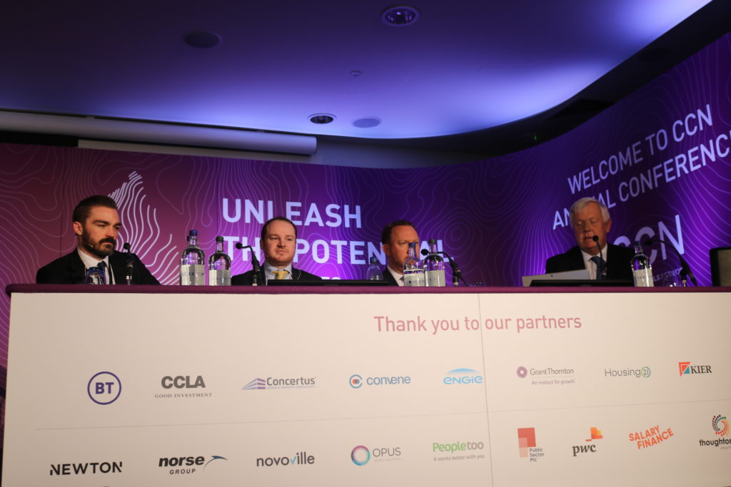Four men seated at conference panel under purple lighting with sponsor logos below