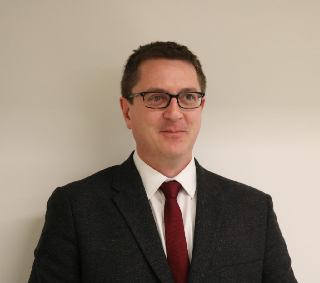 Professional headshot of man wearing glasses, dark suit, white shirt, and burgundy tie.