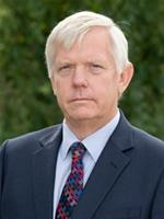 Professional headshot of an older man with white hair wearing a dark blazer and tie.