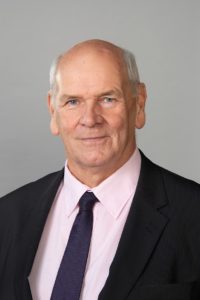 Older man with balding hair wearing pink shirt, navy tie and dark jacket