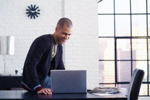 Professional man in dark shirt working at laptop at modern office desk with window view.