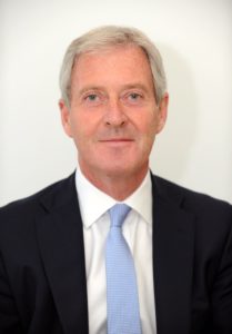 Middle-aged man with gray hair wearing dark blazer and light blue tie portrait.