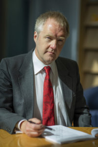 Mature man in gray blazer and red tie sitting at table, writing on document indoors.