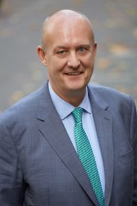 Middle-aged man in blue suit and teal tie smiling at camera against blurred background