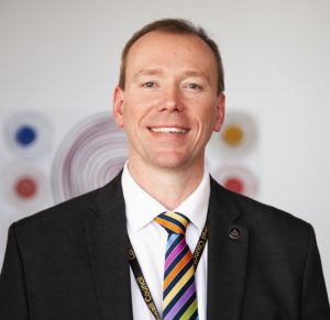 Professional headshot of smiling man in black suit with colorful striped tie at event