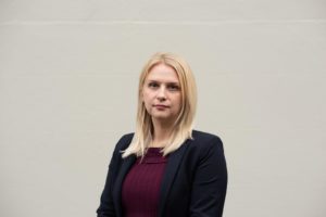Woman with blonde hair wearing dark blazer and maroon top against gray background