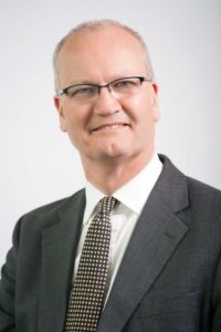 Professional headshot of middle-aged man wearing glasses and dark business suit.