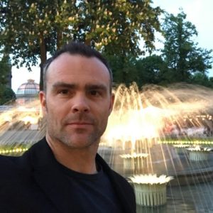 Man in black shirt poses outdoors with illuminated building and trees background