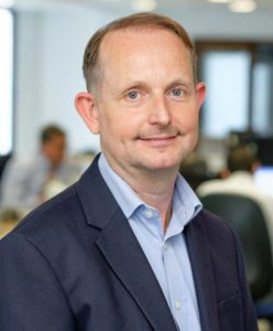 Professional man in dark blazer and light blue shirt smiling at camera indoors.