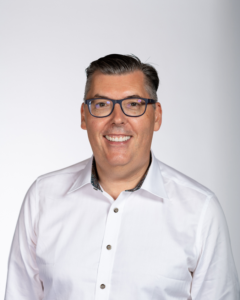 Man wearing glasses and white collared shirt smiling at camera on gray background
