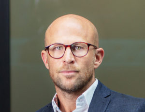 Bald man wearing glasses and blue blazer against green background