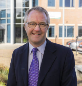 Middle-aged man wearing glasses, dark blazer, and purple tie outdoors.