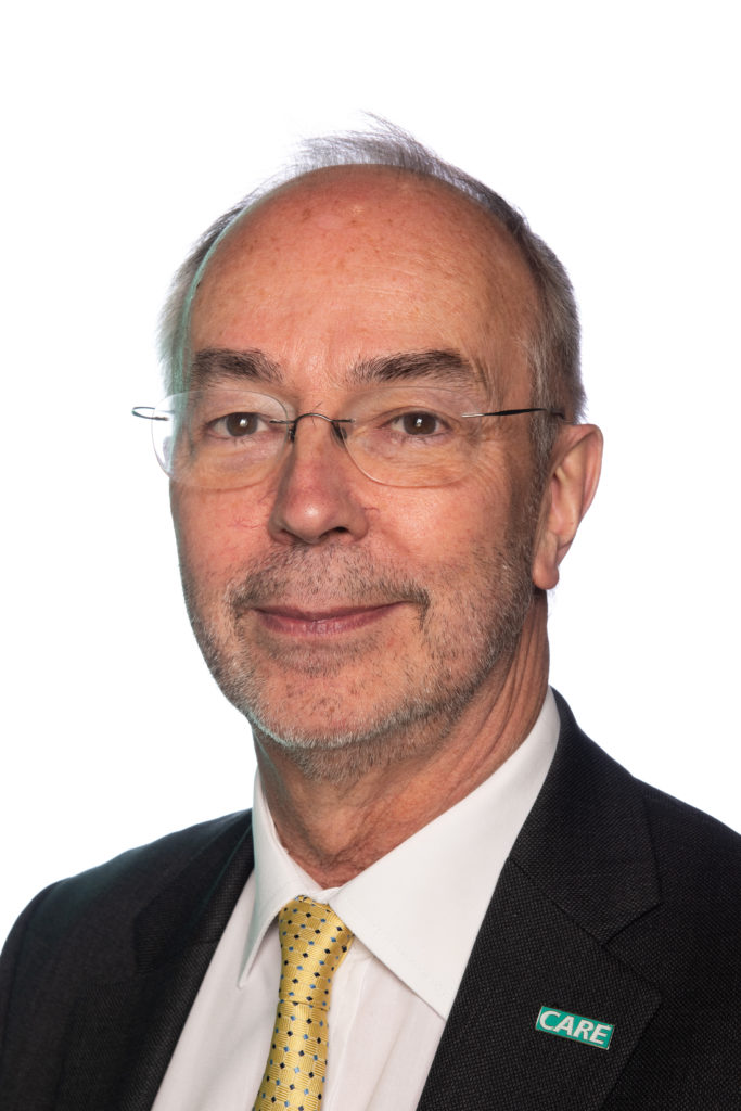 Professional headshot of an older man wearing glasses, dark jacket, white shirt, and yellow tie.