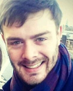 Smiling young man with beard and dark hair wearing dark jacket indoors