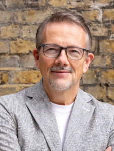 Man wearing glasses and gray blazer smiling against brick wall background
