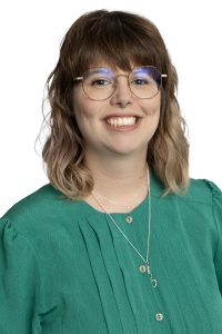 Person with shoulder-length brown hair, blue-framed glasses, and teal green shirt smiling.
