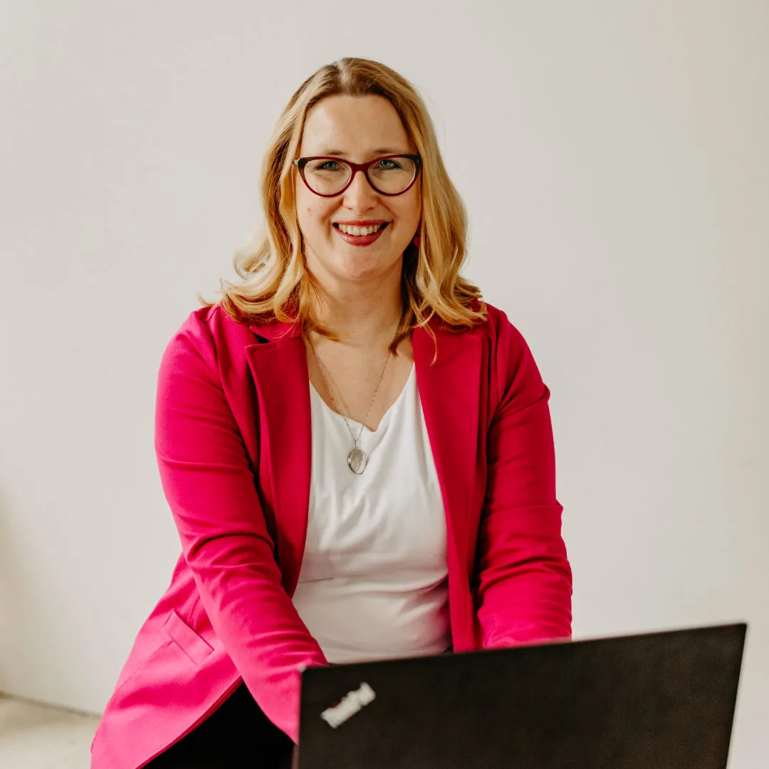 Smiling Louisa Hoffschult with glasses in pink blazer using a black ThinkPad laptop against white background.