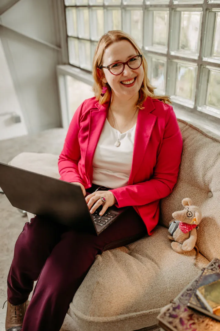 Smiling Louisa Hoffschult in glasses and pink blazer working on a laptop, seated on beige couch next to a stuffed animal named Liesel Leseratte.