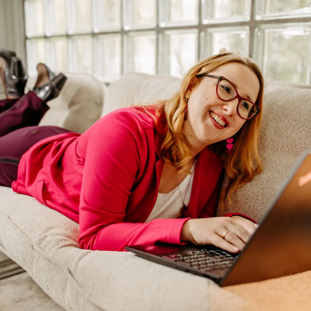 Smiling Louisa Hoffschult in pink jacket and glasses lying on a couch using a laptop.