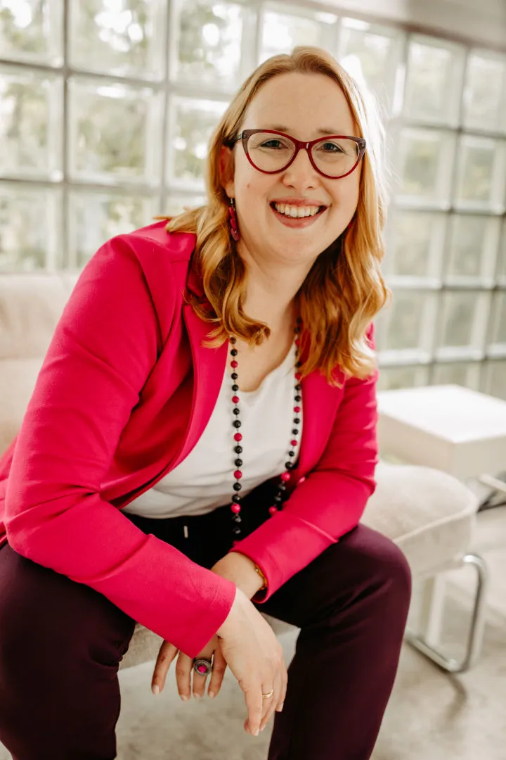 Smiling Louisa Hoffschult with light hair, red glasses, and a bright pink blazer sitting indoors on a beige chair.