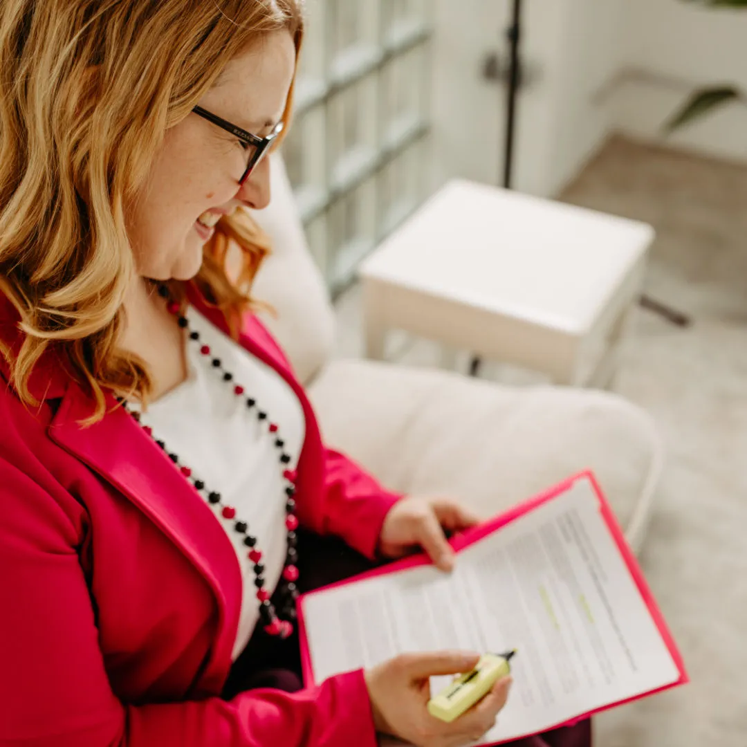 Smiling Louisa Hoffschult in glasses and pink blazer highlighting text on a clipboard while seated on a couch.