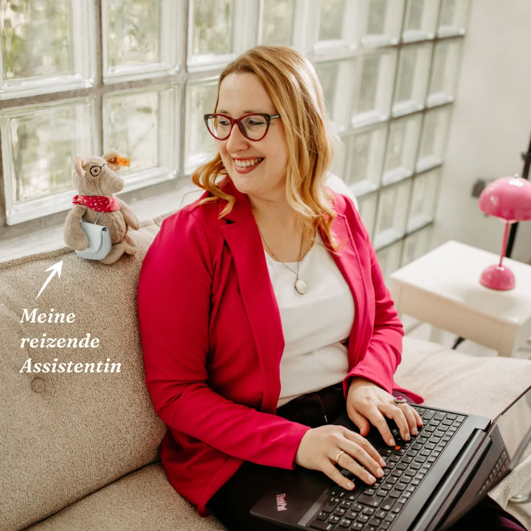 Smiling Louisa Hoffschult in pink blazer typing on laptop with stuffed animal Liesel Leseratte and German text on couch beside her.