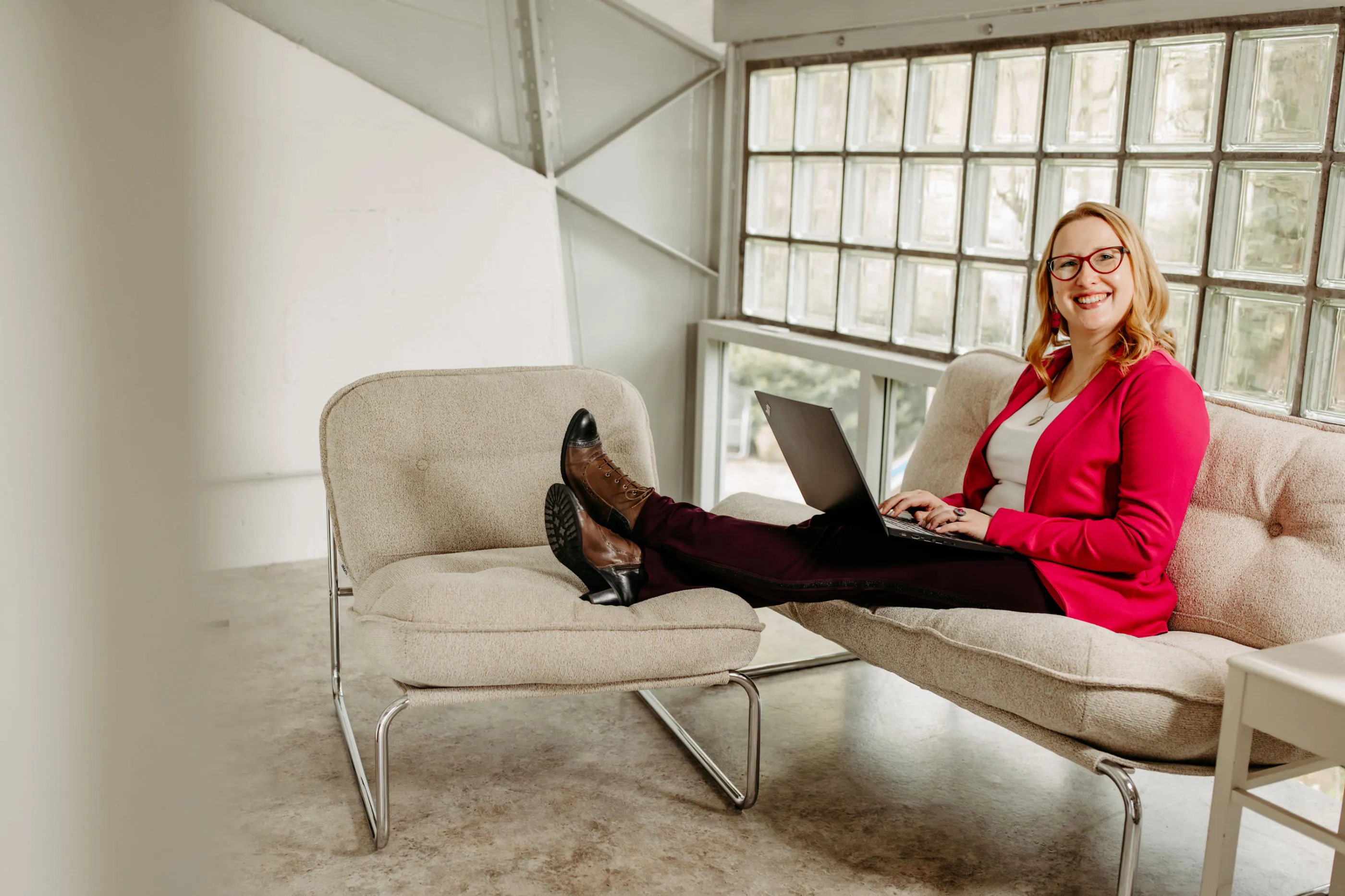 Smiling Louisa Hoffschult in pink blazer sitting on beige sofa with laptop and legs on an armchair.