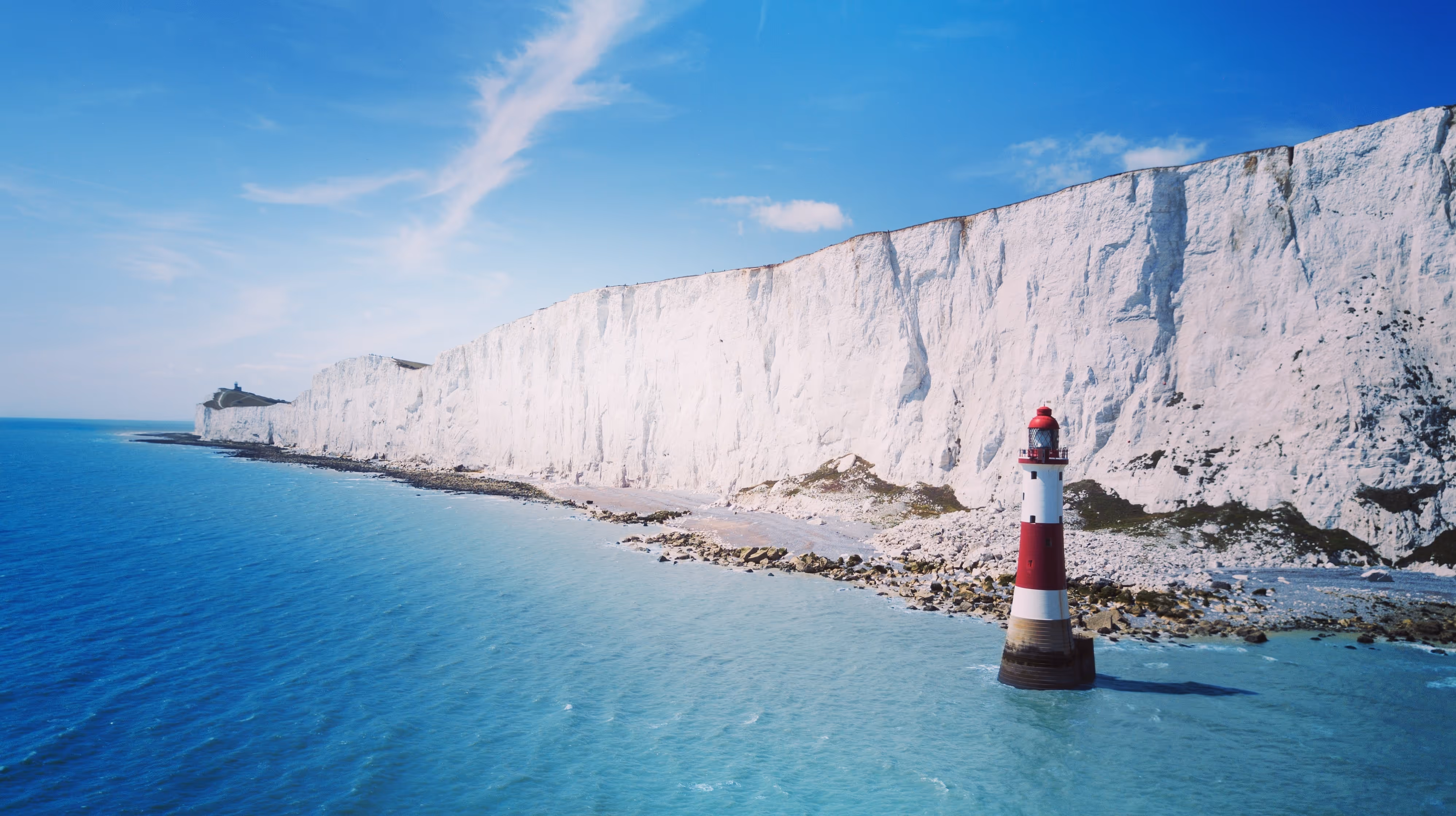 A lighthouse stands in the sea near tall white cliffs under a clear blue sky.