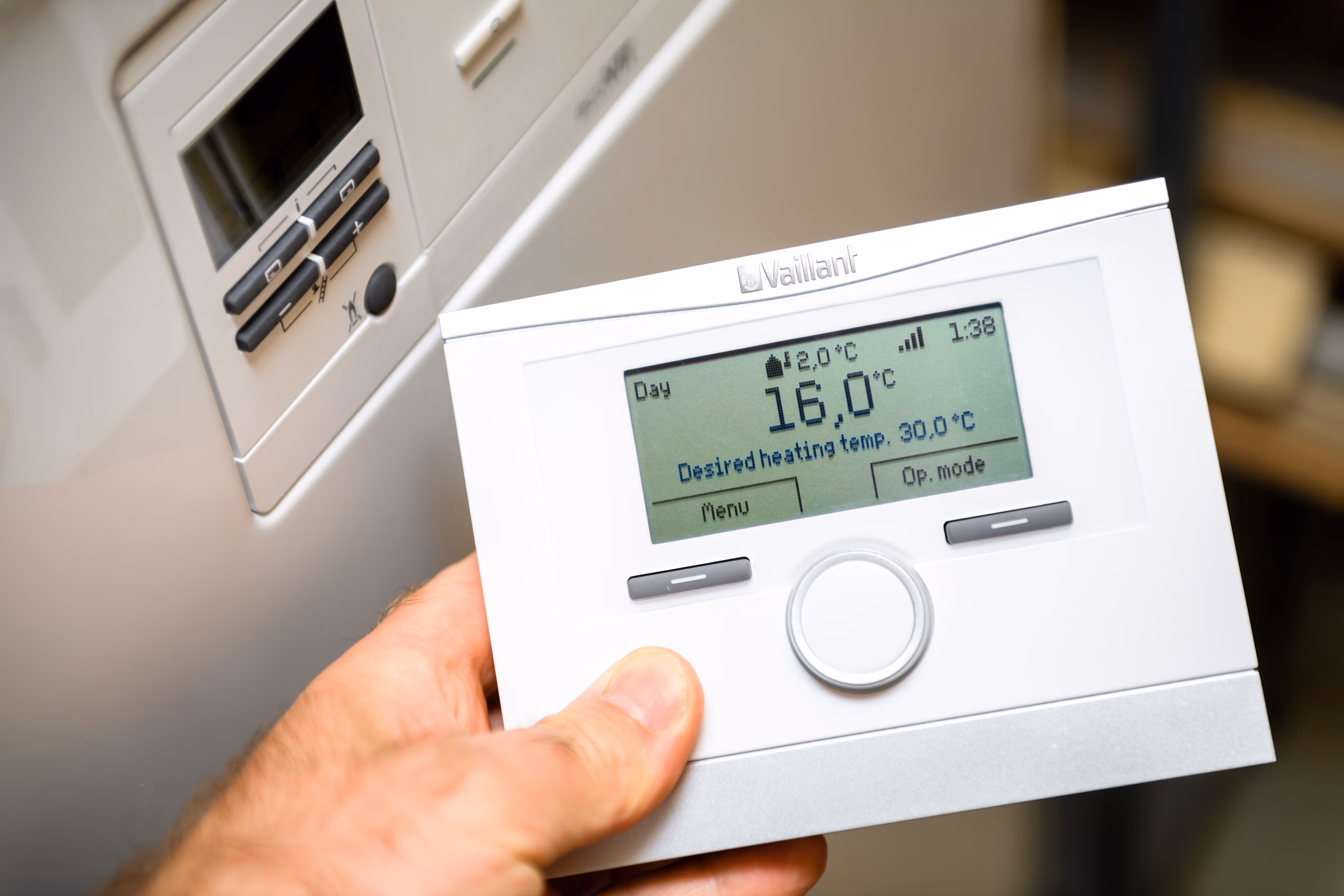 A hand holds a digital thermostat displaying the temperature settings for indoor heating.