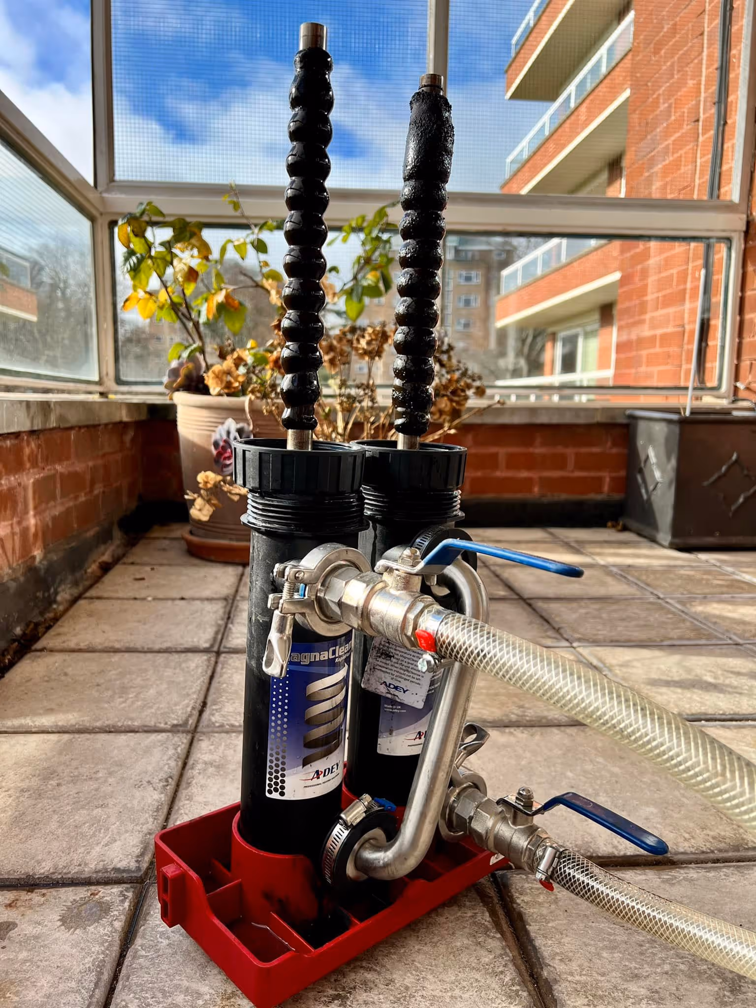 Two filter canisters connected with hoses on a patio, with plants and a building in the background.