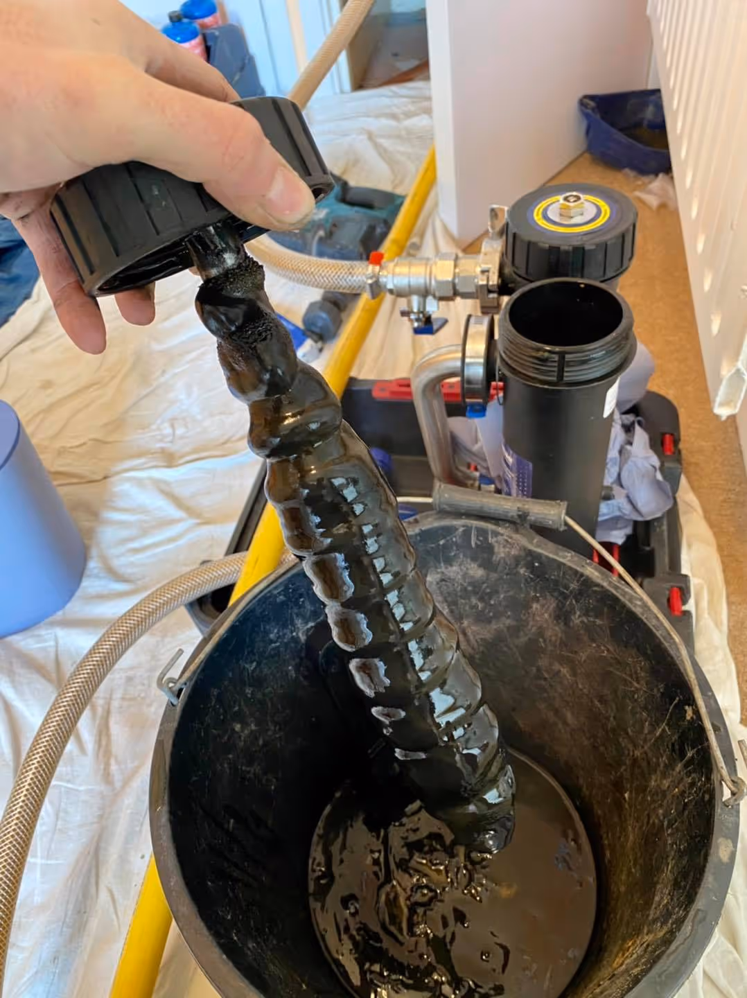 A person holds a thick, black, sludge-covered object over a bucket, possibly cleaning or maintaining equipment.