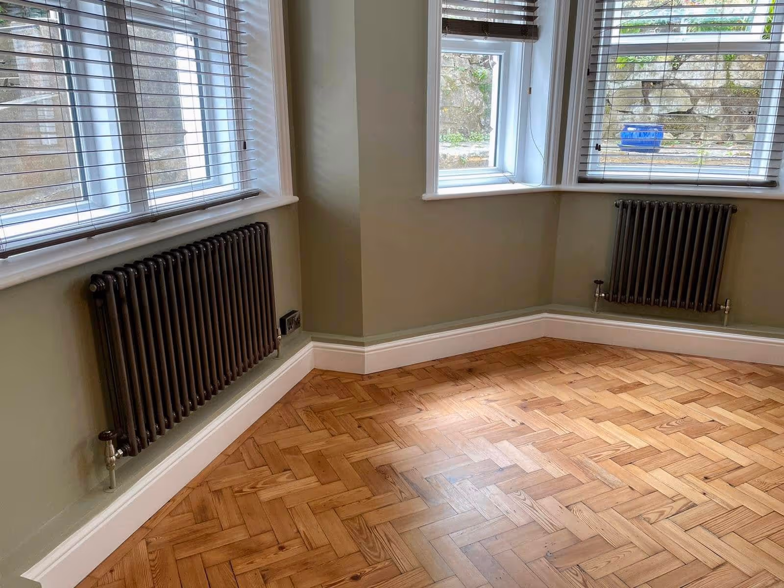 Empty room with wooden herringbone floor, two radiators, and large windows with blinds.