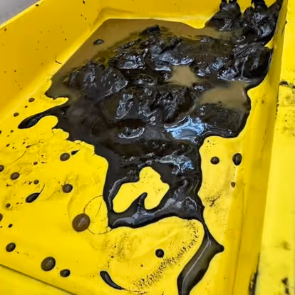 Thick, black oil sludge in a yellow container, with small puddles and splatters around it.