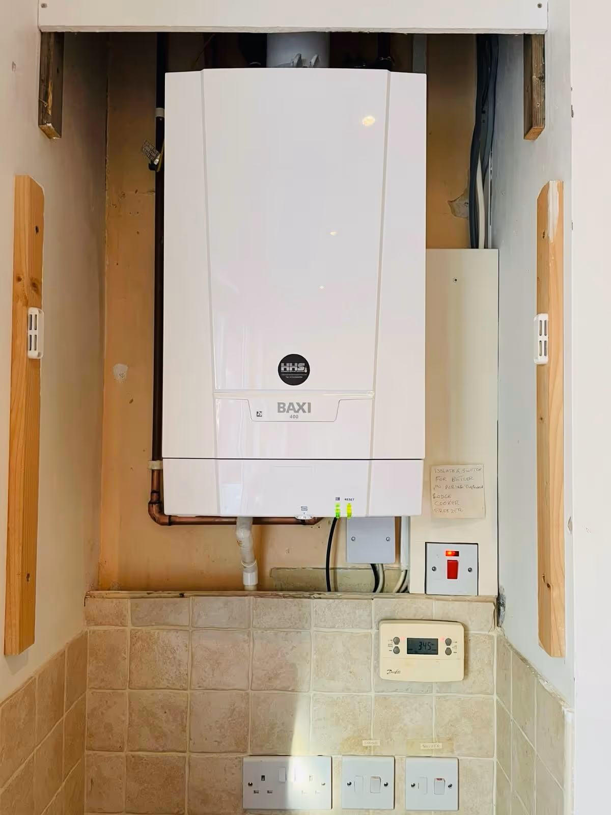 Wall-mounted white boiler with control panel and pipes, above light-colored tiled surface and electrical outlets.