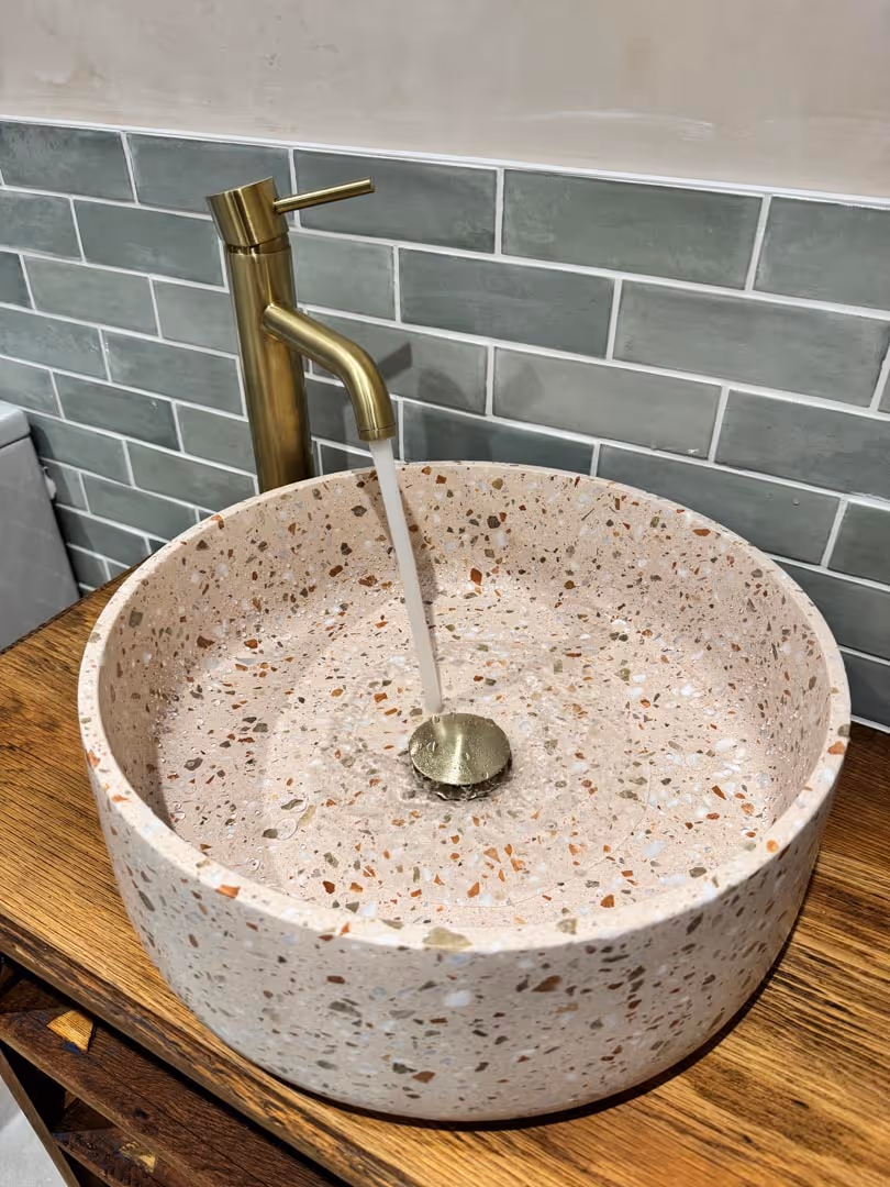 Round stone sink with flowing water, gold faucet, and gray tile backsplash on a wooden countertop.