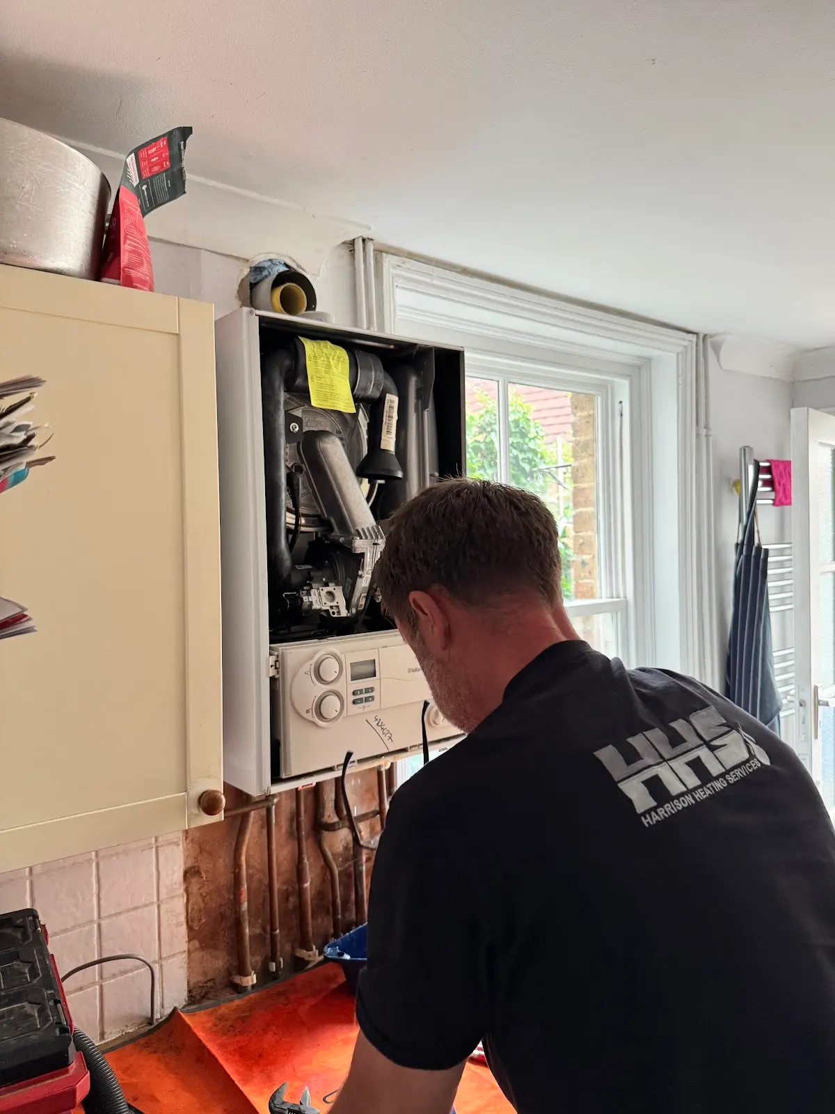 A man is inspecting a wall-mounted boiler with an open cover in a room.