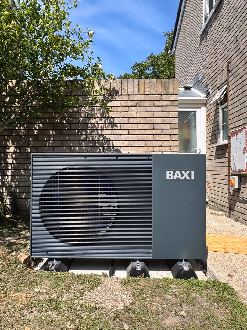 A large Baxi heat pump is installed outside a brick house on a concrete surface.