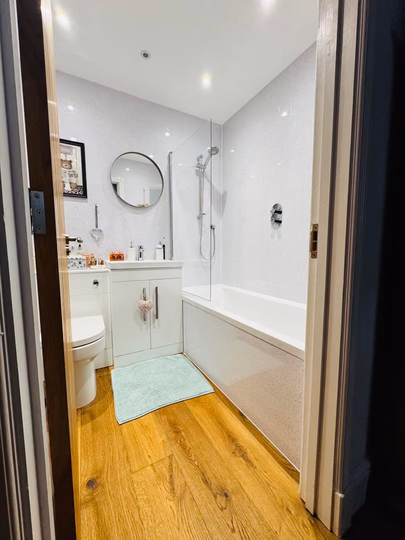 Bathroom with a bathtub, shower, sink, and round mirror. Light wooden floor and a bath mat.