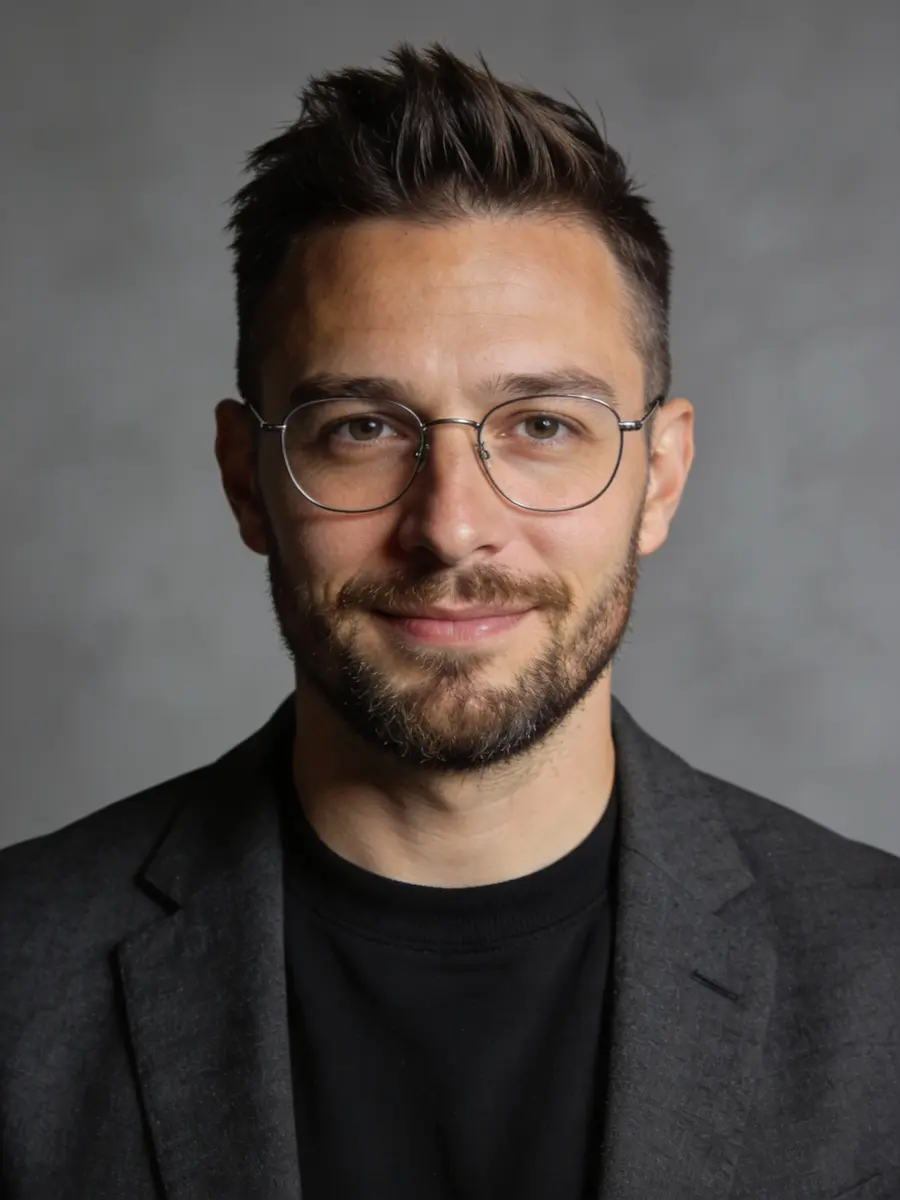 Smiling man with short dark hair, glasses, beard, wearing a black shirt and dark blazer, gray background.