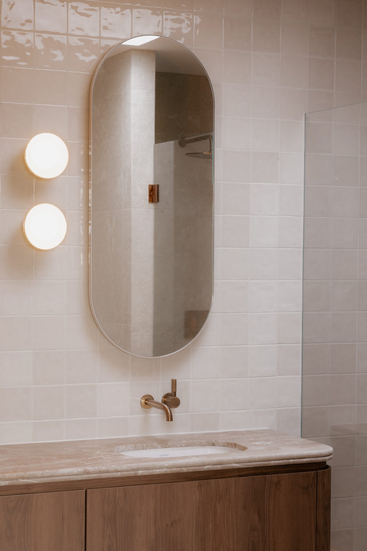 Toorak House bathroom with cream tiles, oval capsule mirror, brass tapware, stone benchtop and walnut vanity
