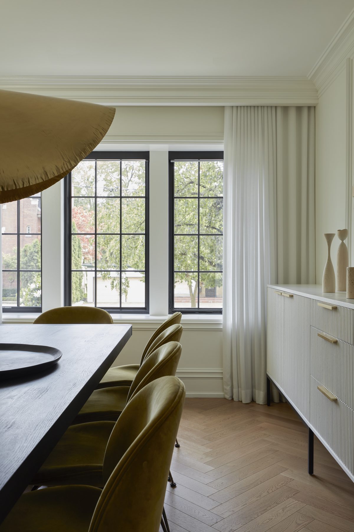 Midtown House dining room with brass pendant, olive velvet chairs, dark wood table and black-framed grid windows