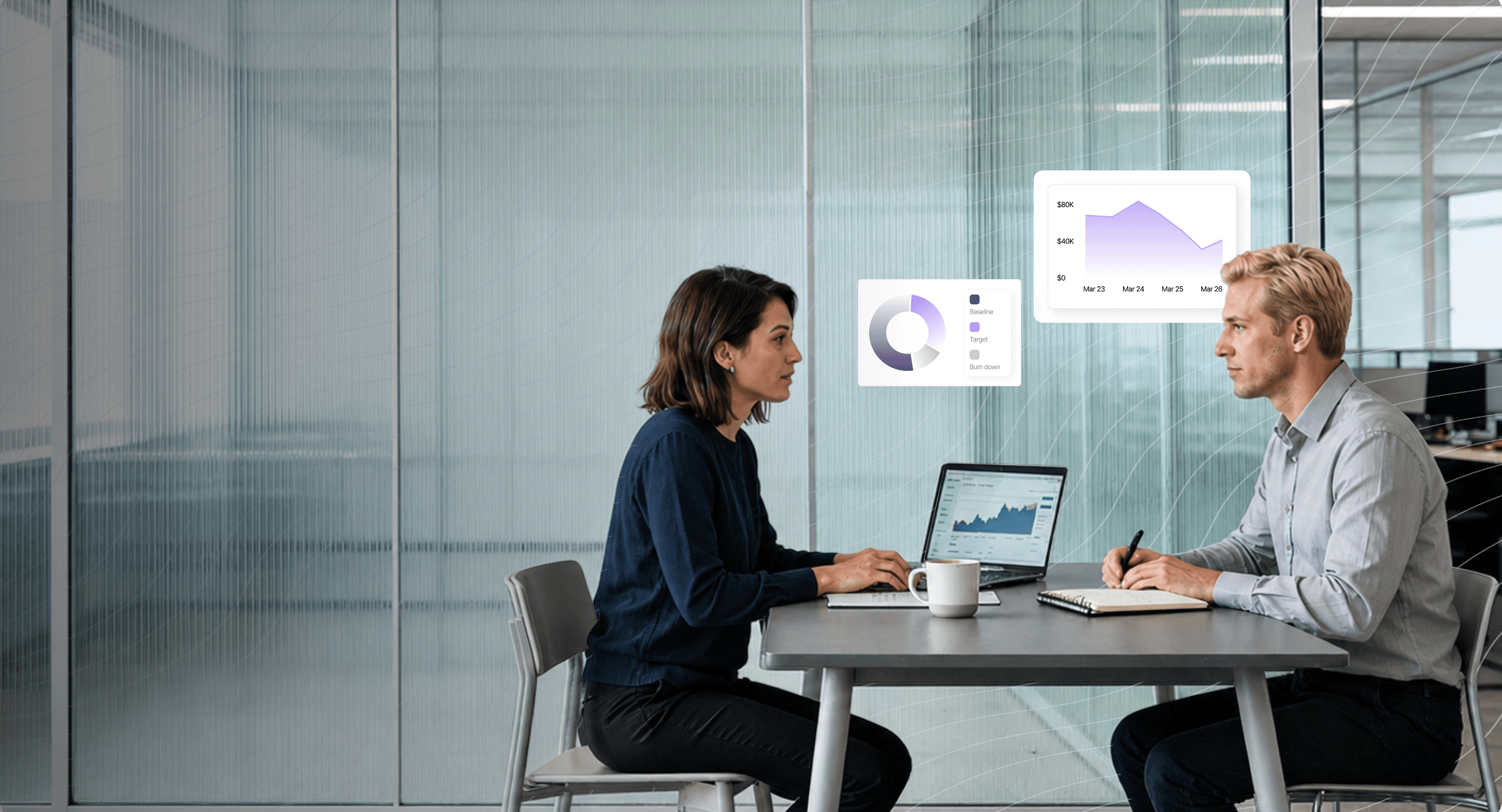 Two coworkers sit across a table in an office with a laptop showing charts and graphs between them.
