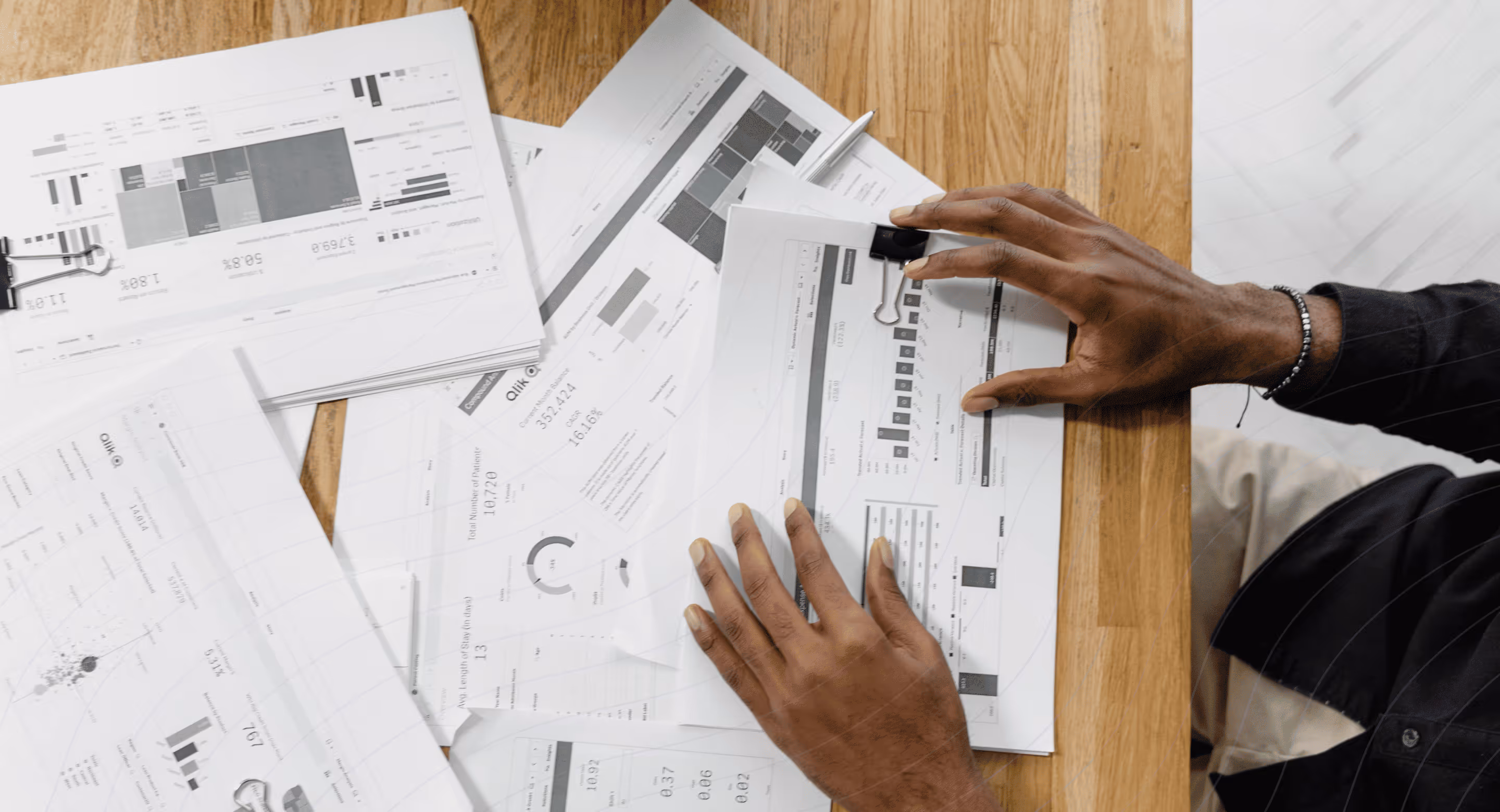 Hands organizing printed reports and charts on a wooden table with a pen nearby.