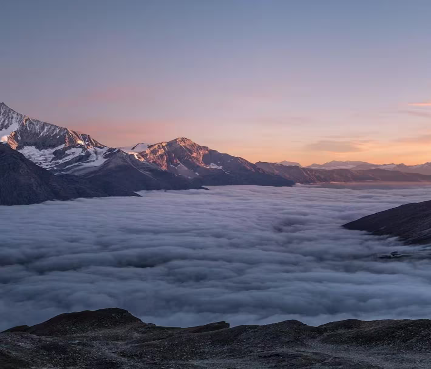 Mountain range with snow-capped peaks above thick clouds at sunset.