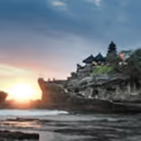 Sunset over rocky coastline with a temple structure on a cliff surrounded by water and sky.
