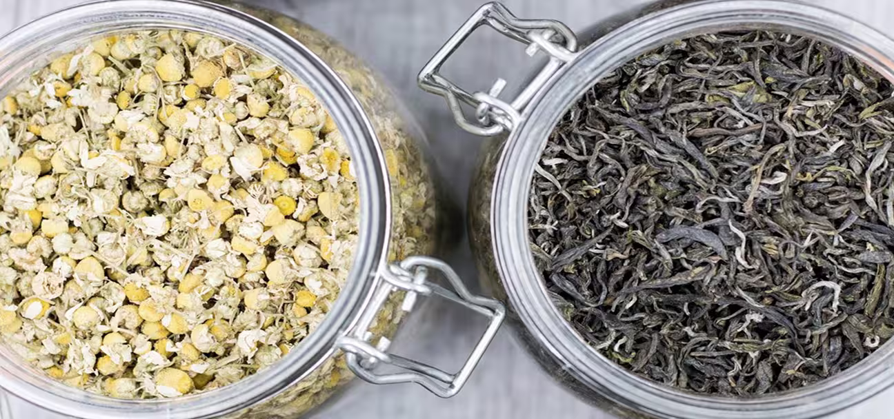 Top view of two glass jars filled with dried chamomile flowers and loose black tea leaves.