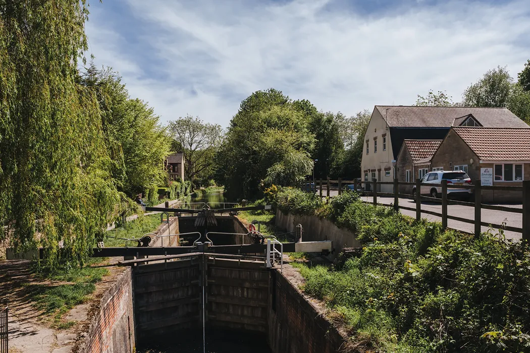 A canal lock surrounded by greenery, trees, and buildings on a partly cloudy day.
