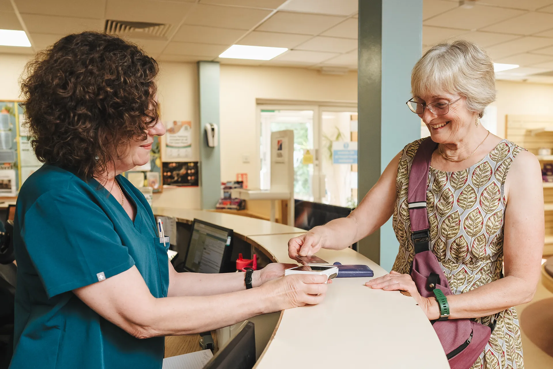 Smiling older woman paying with card held over a terminal by a woman in medical scrubs at reception.