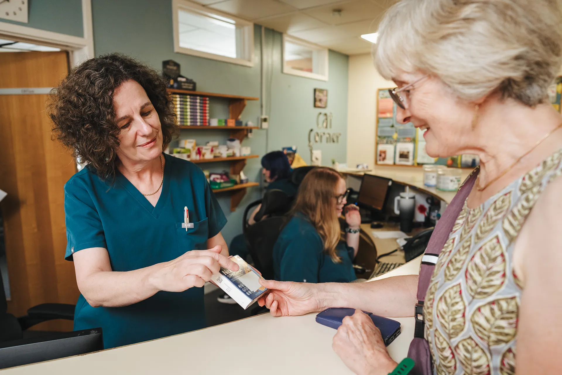 Veterinary staff member in scrubs handing a product to an older woman at a clinic reception desk.